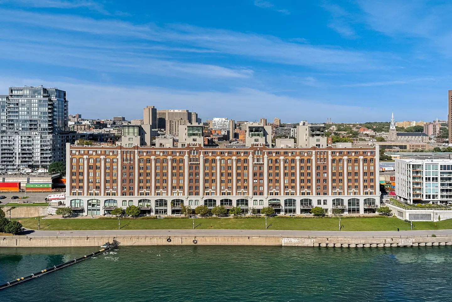 Exterior view of a brick apartment building with many windows, next to a river and a city skyline under a blue sky.