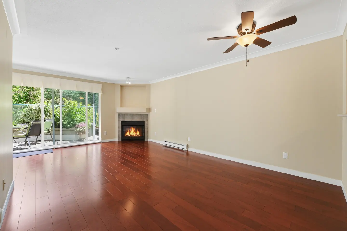 Bright living room with hardwood floors, beige walls, and a fireplace. Sliding glass doors lead to a patio with greenery. Ceiling fan overhead.