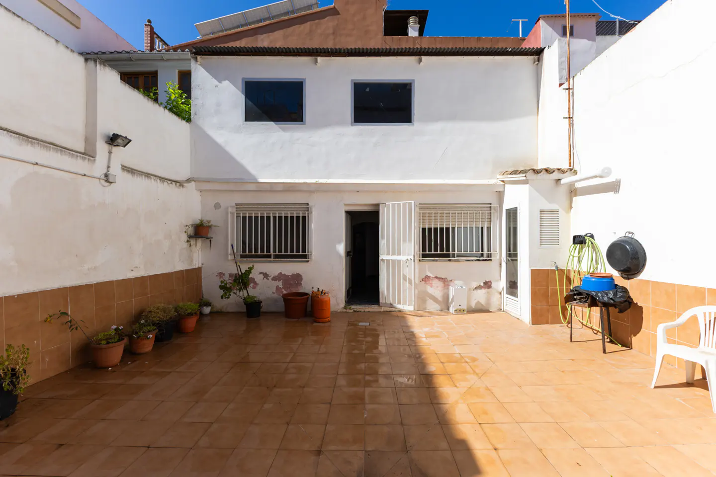 A sunny backyard with a tiled floor, white walls, and a two-story white house with barred windows and an open door.