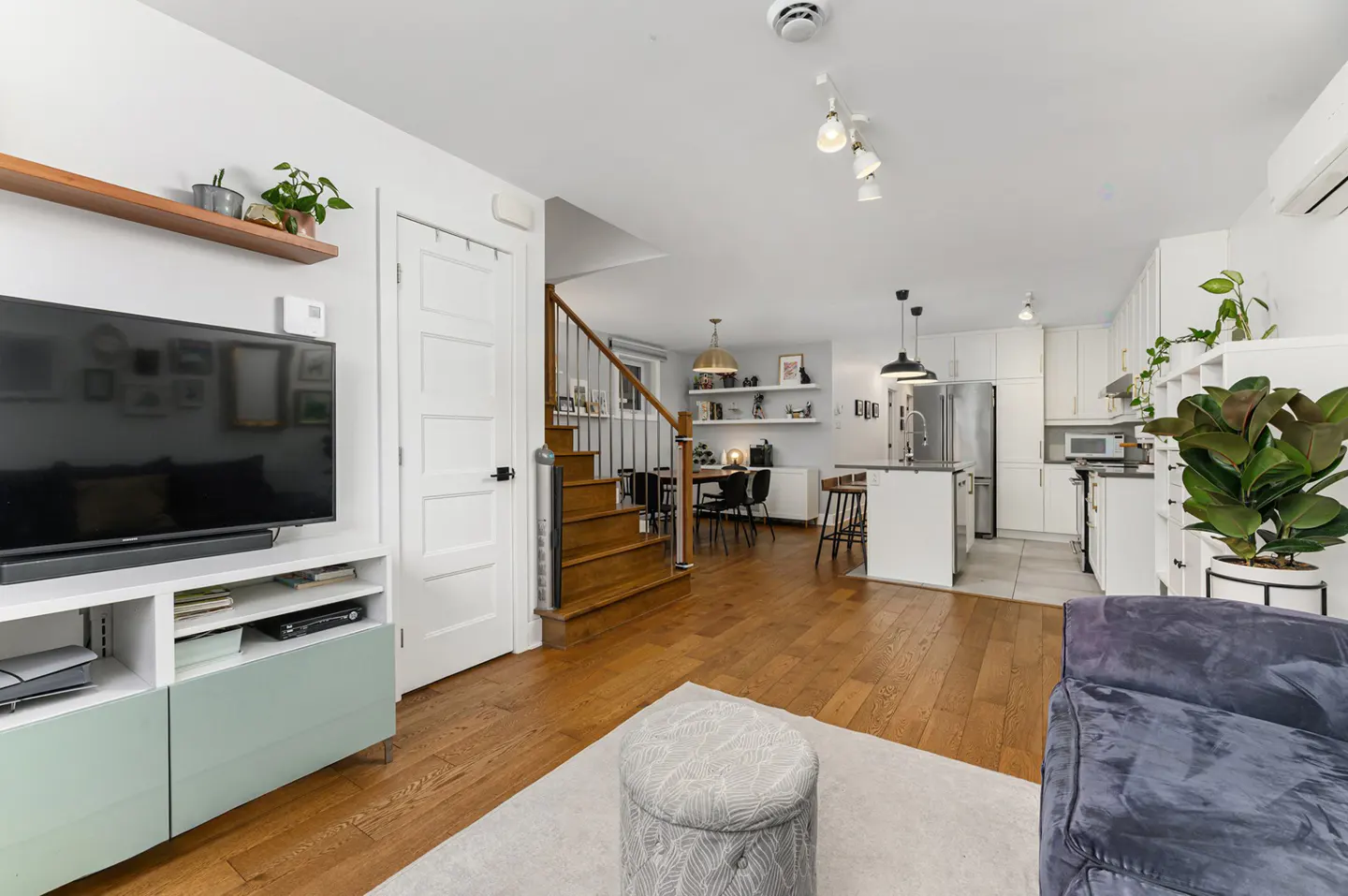 Open-concept living space with hardwood floors, white walls, and a staircase. A TV sits on a light green cabinet, and a kitchen is visible in the background.