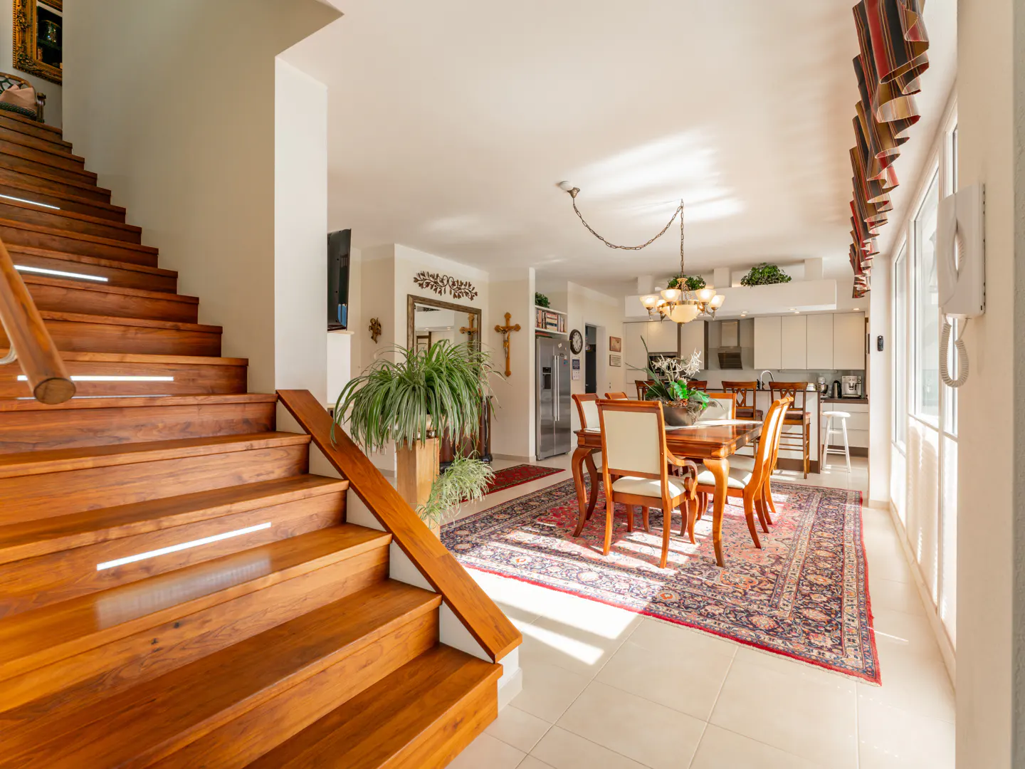 Bright, open home interior with wood stairs, dining table, and kitchen. A patterned rug sits under the dining set. Sunlight streams through windows.