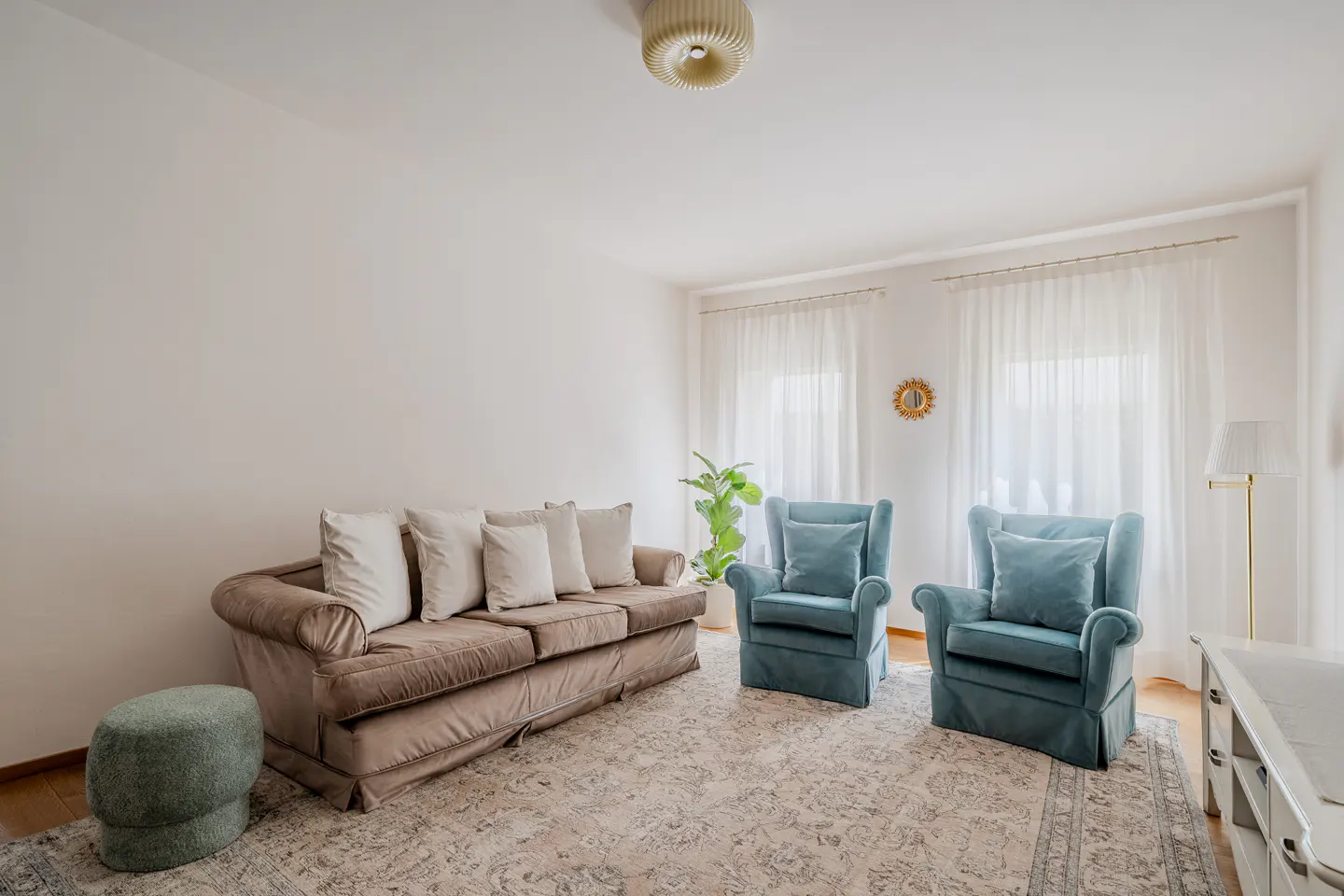 Living room with a brown sofa, two blue armchairs, and a patterned rug. White walls and sheer curtains create a bright, airy space.