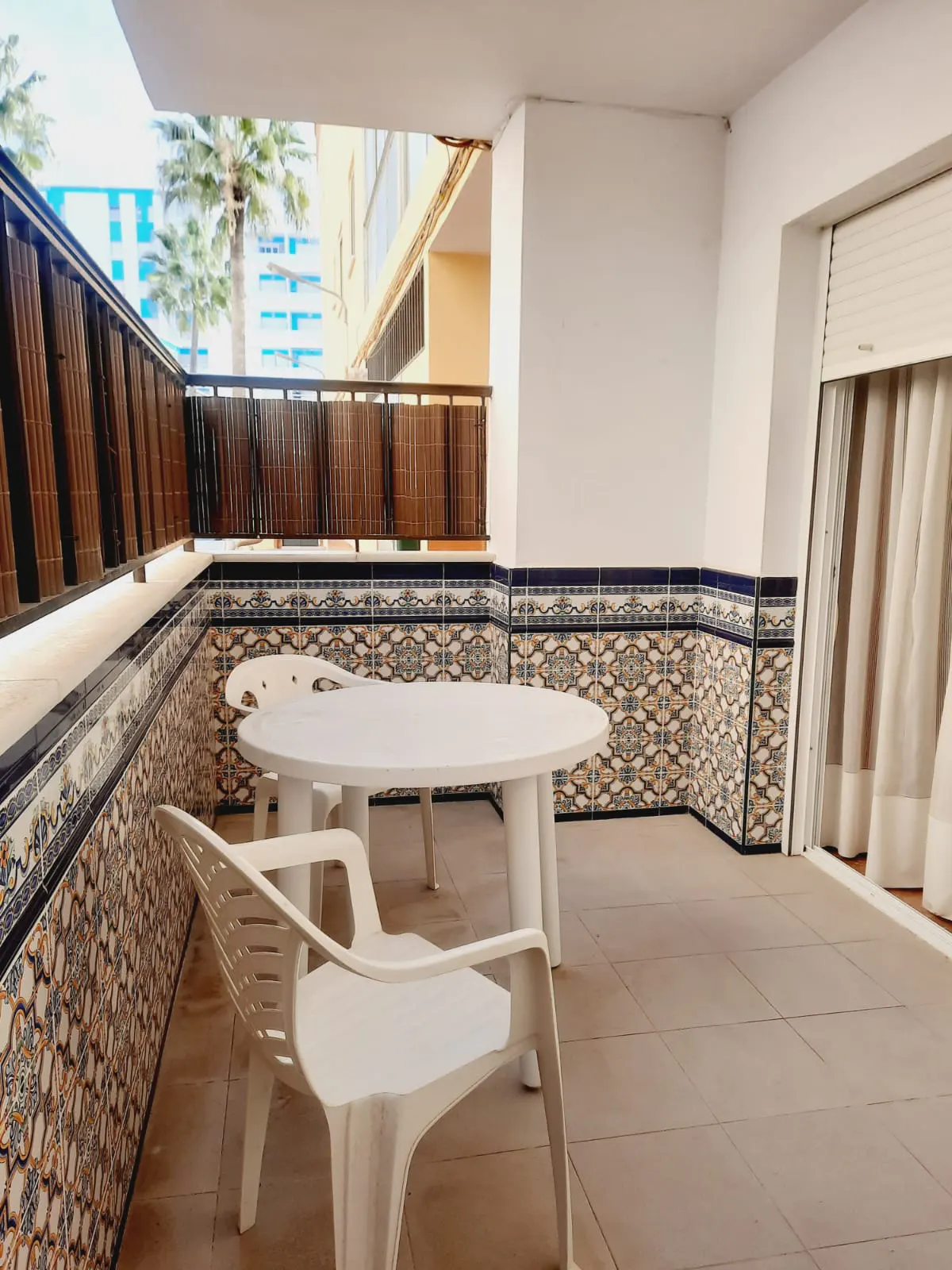 A balcony with a white table and chairs, tiled walls, and a view of palm trees.