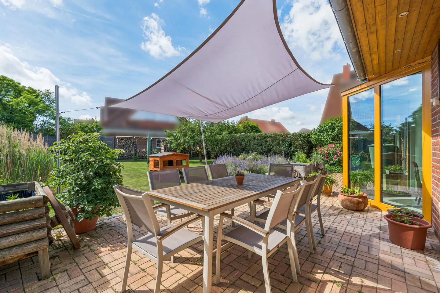 A sunny patio with a wooden dining set under a white shade sail, overlooking a green backyard with a small wooden shed.