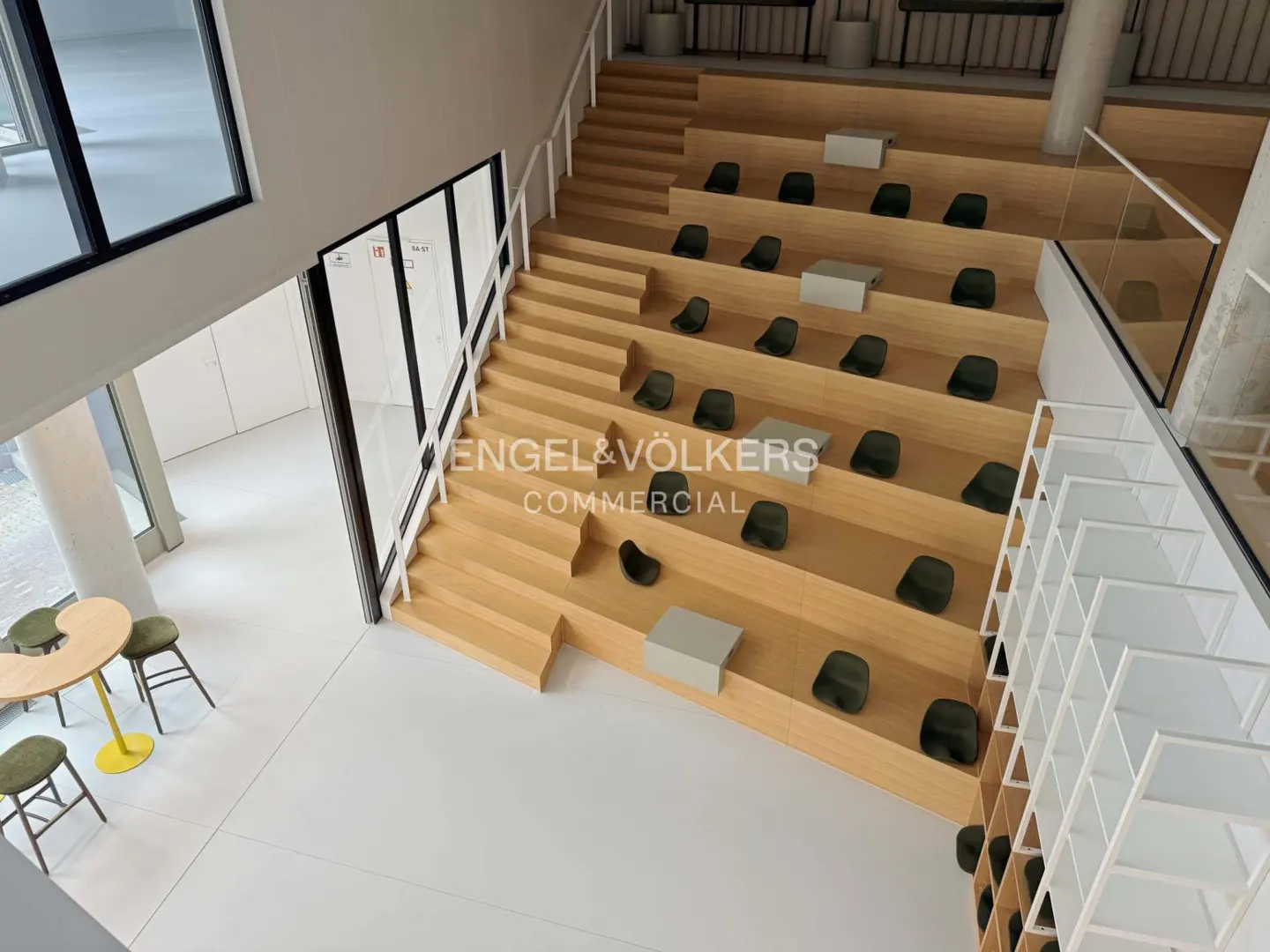 Interior view of a tiered seating area with wooden steps and dark green chairs, plus a table and chairs on a white floor.