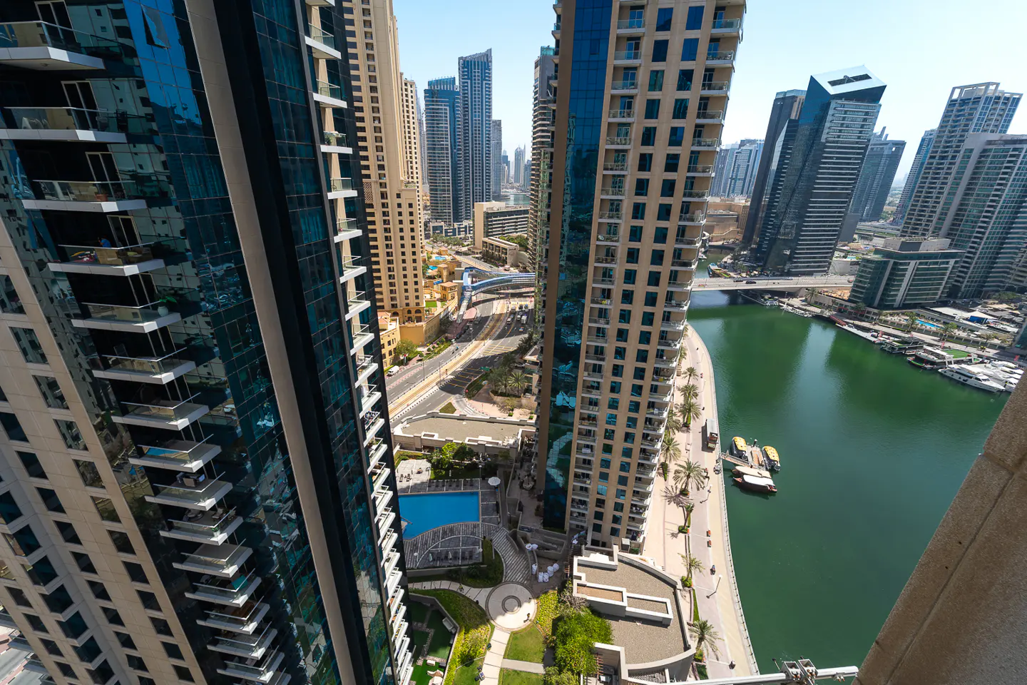 View from a high-rise balcony of Dubai Marina, showing skyscrapers, a green canal, and a pool area below.
