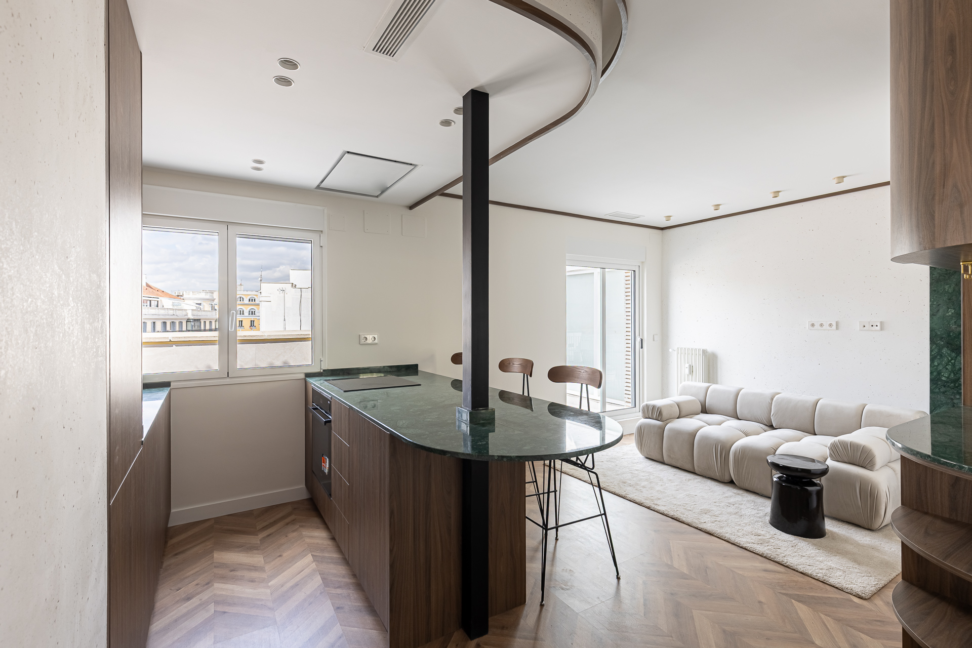 Open-concept apartment with a kitchen island, wood floors, and a white sofa on a rug. A window overlooks a city.