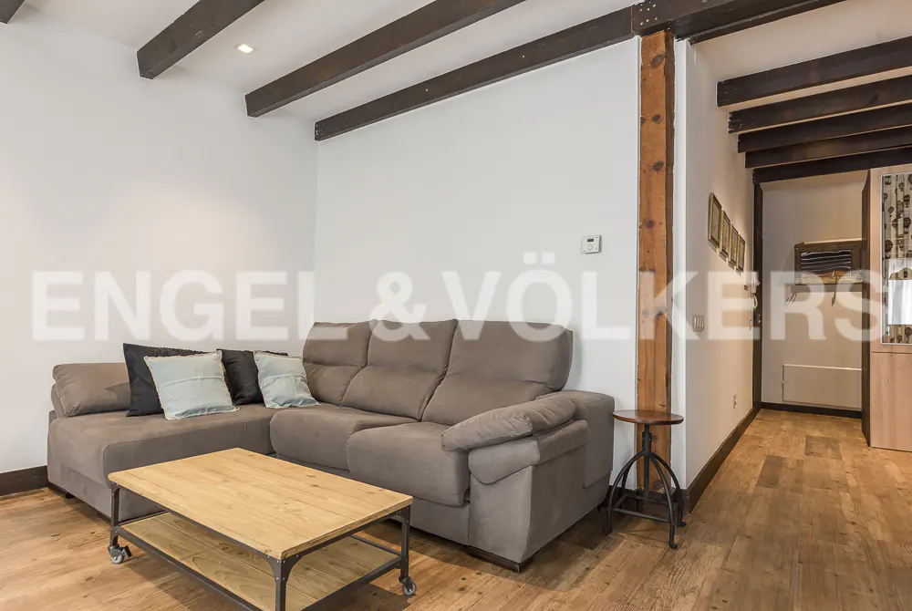 Living room with brown sofa, wooden coffee table, and exposed beams. Hallway visible in the background.