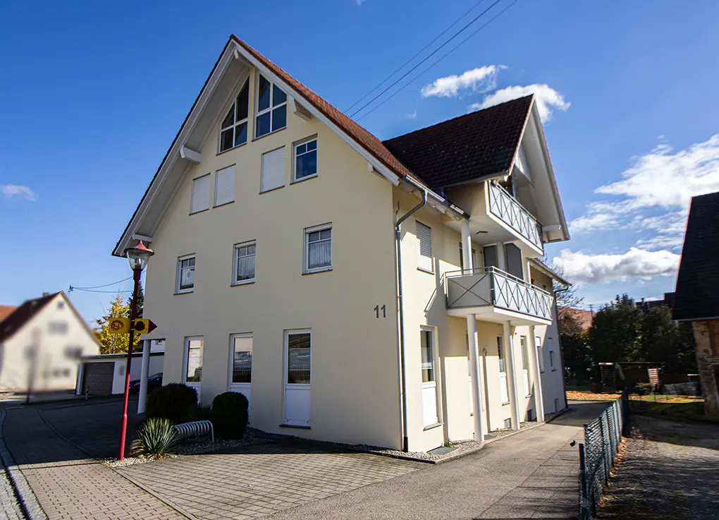 Three-story cream-colored house with a brown roof, balconies, and the number 11 on the side, set under a blue sky.