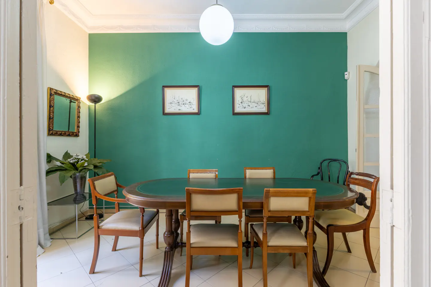 A dining room with a green wall, an oval table with six chairs, and two framed pictures.
