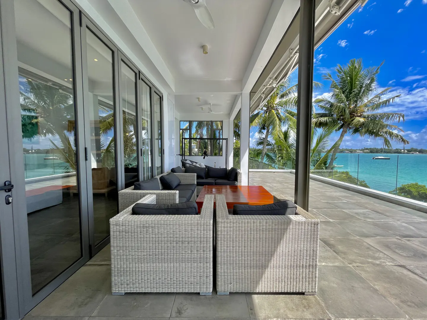 Outdoor patio with wicker furniture, gray cushions, and a wood table. Ocean view with palm trees and blue sky in the background.