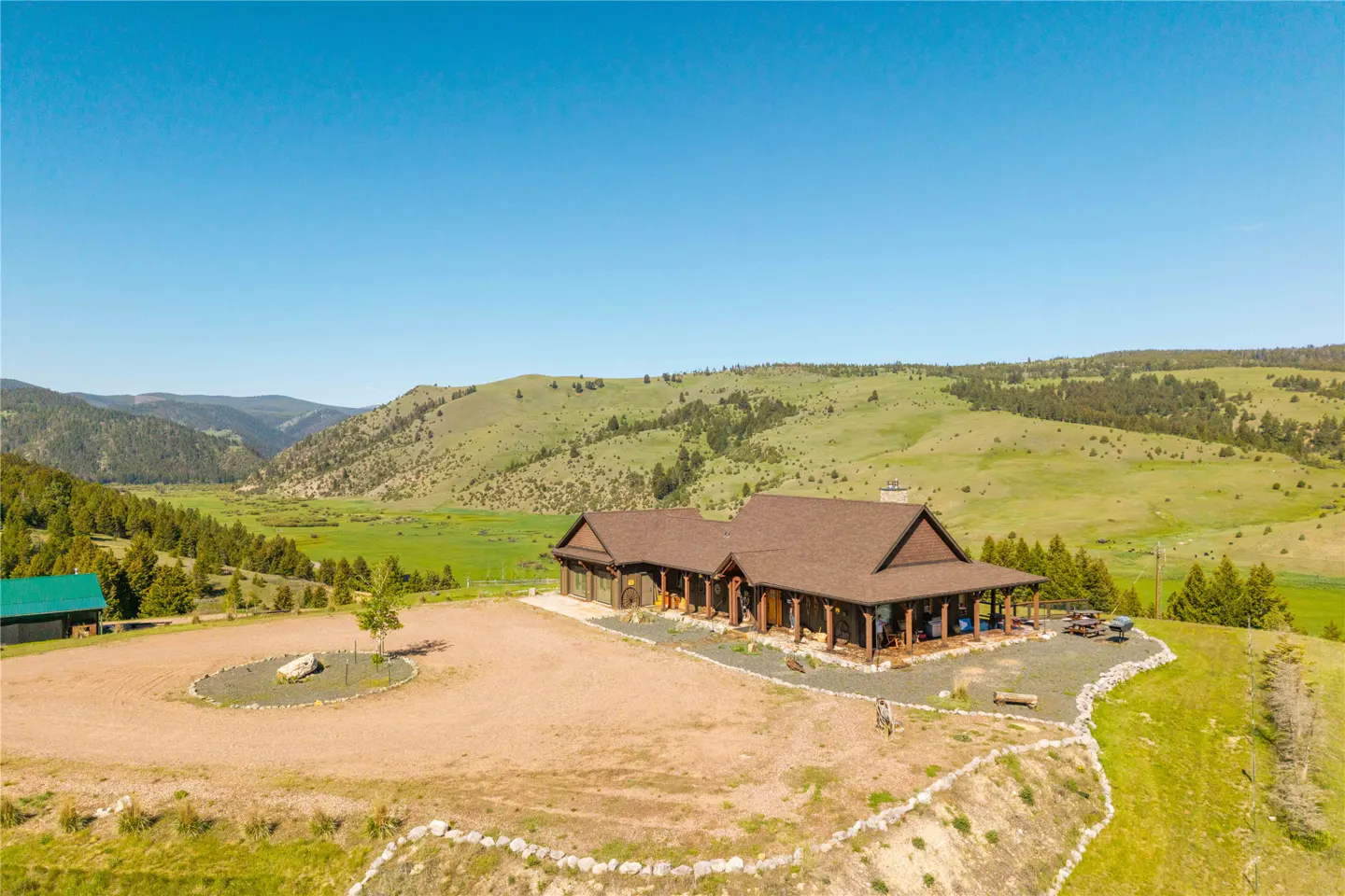 Aerial view of a brown ranch-style home with a wrap-around porch, set in a vast, green mountain valley under a blue sky.