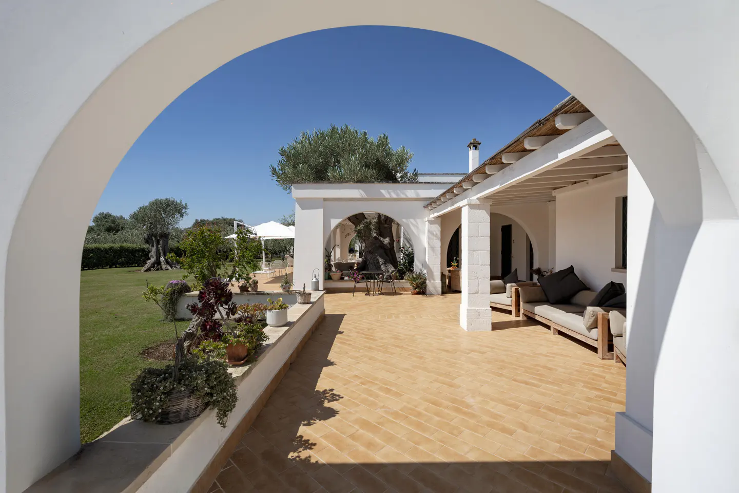Exterior view of a white villa with arched doorways, a terracotta patio, and green lawn under a clear blue sky.