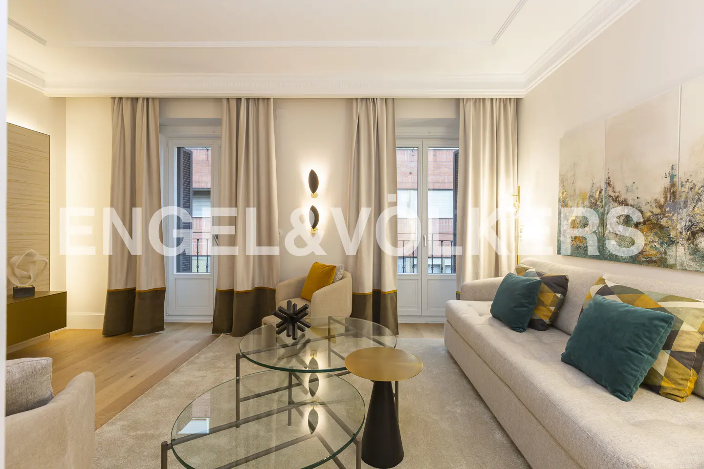 Living room with beige sofa, teal pillows, glass tables, and beige curtains. Artwork hangs on the wall.