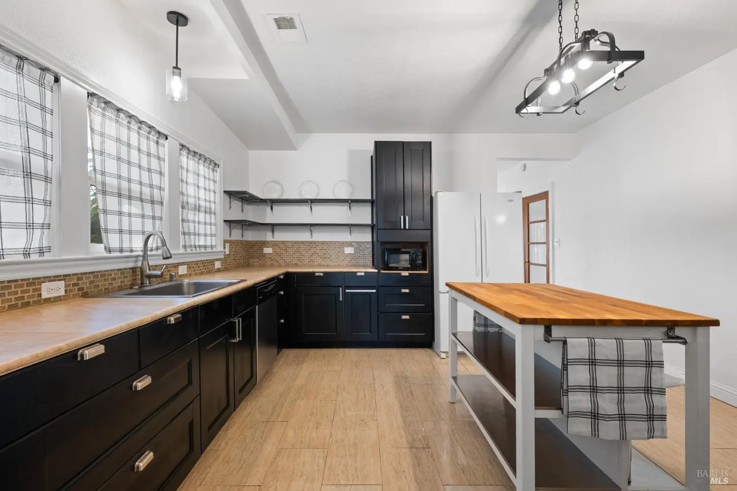 Bright kitchen with black cabinets, tan countertops, and a wood-topped island. Plaid curtains hang over the sink.