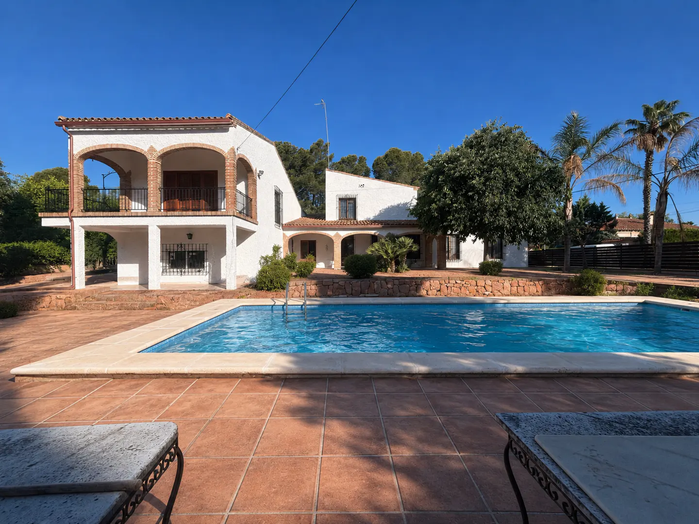 A white two-story house with a pool and a red tile patio under a clear blue sky.
