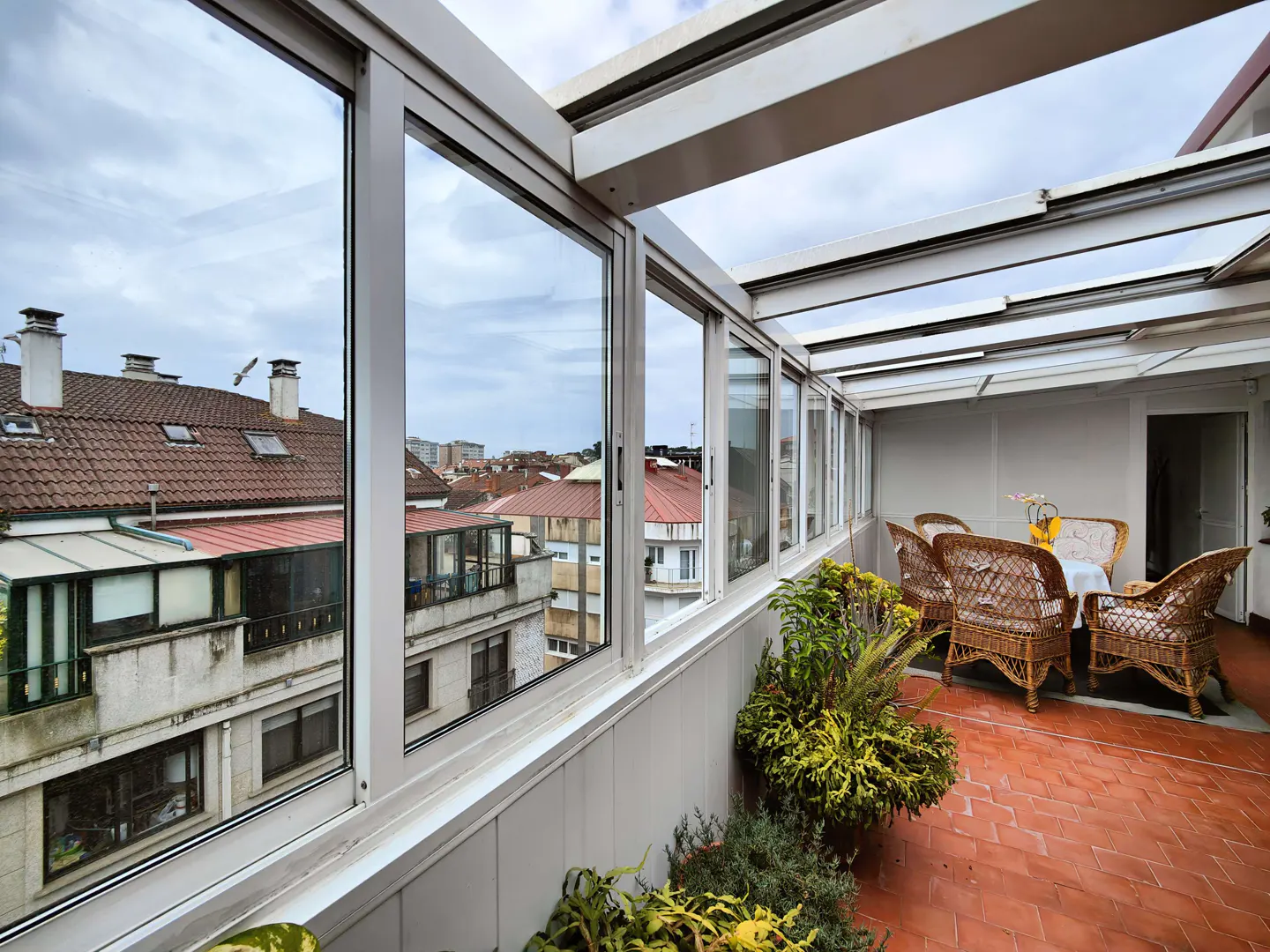 Enclosed patio with wicker furniture, plants, and a view of rooftops through large windows.