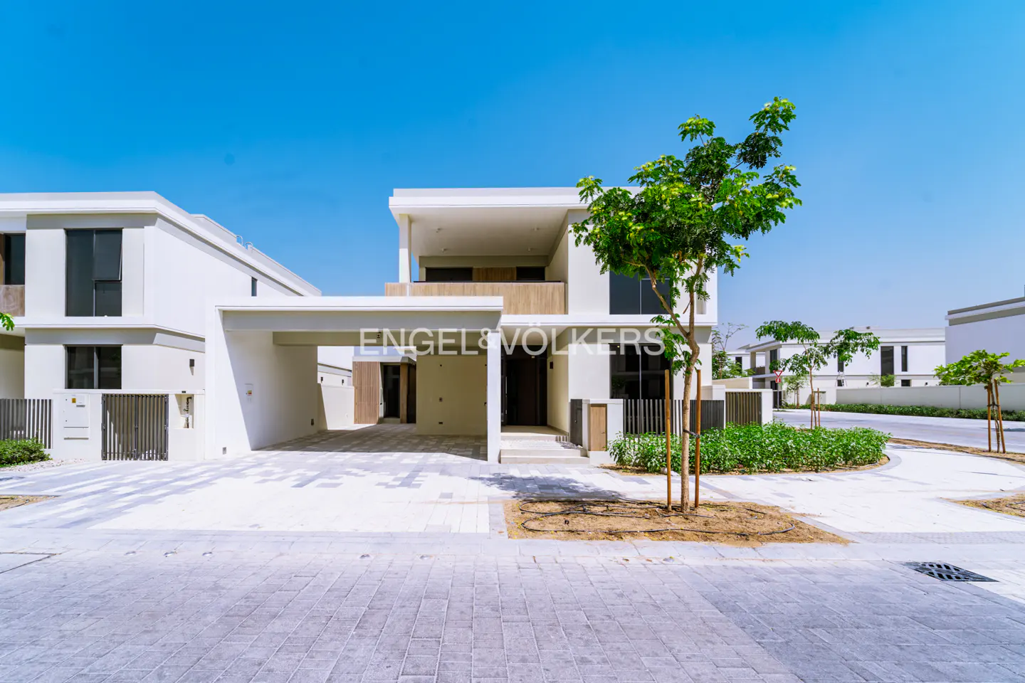 Modern white two-story villa with a covered carport, wood accents, and a small front garden under a clear blue sky.