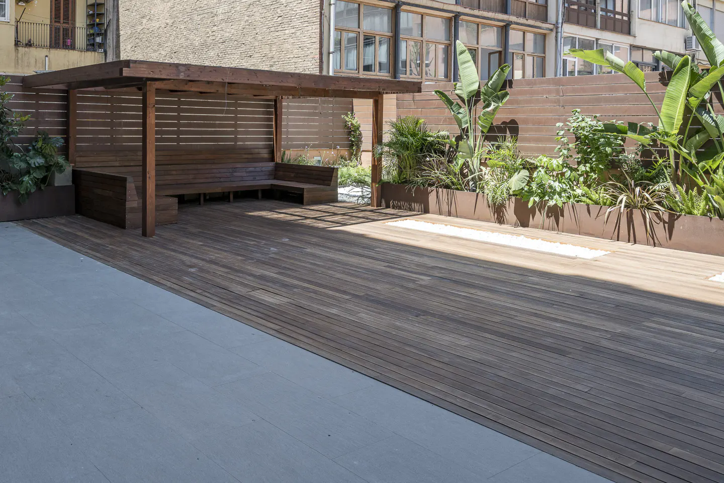 Modern wooden deck with a pergola, built-in bench seating, and lush green plants against a wooden privacy fence.