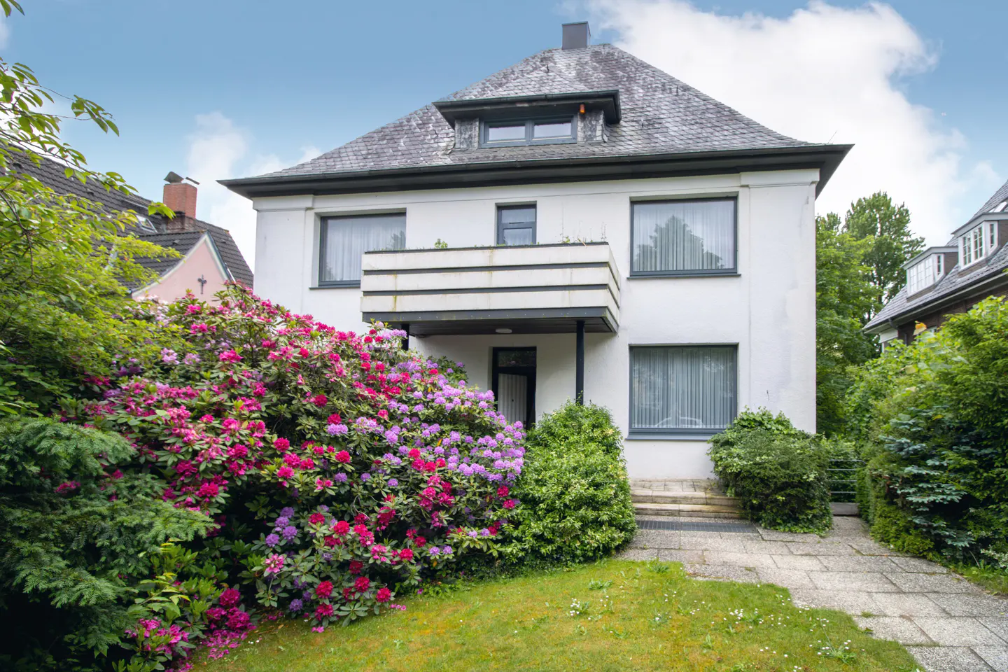 White two-story house with a balcony, slate roof, and a large, vibrant pink and purple rhododendron bush in the front yard.