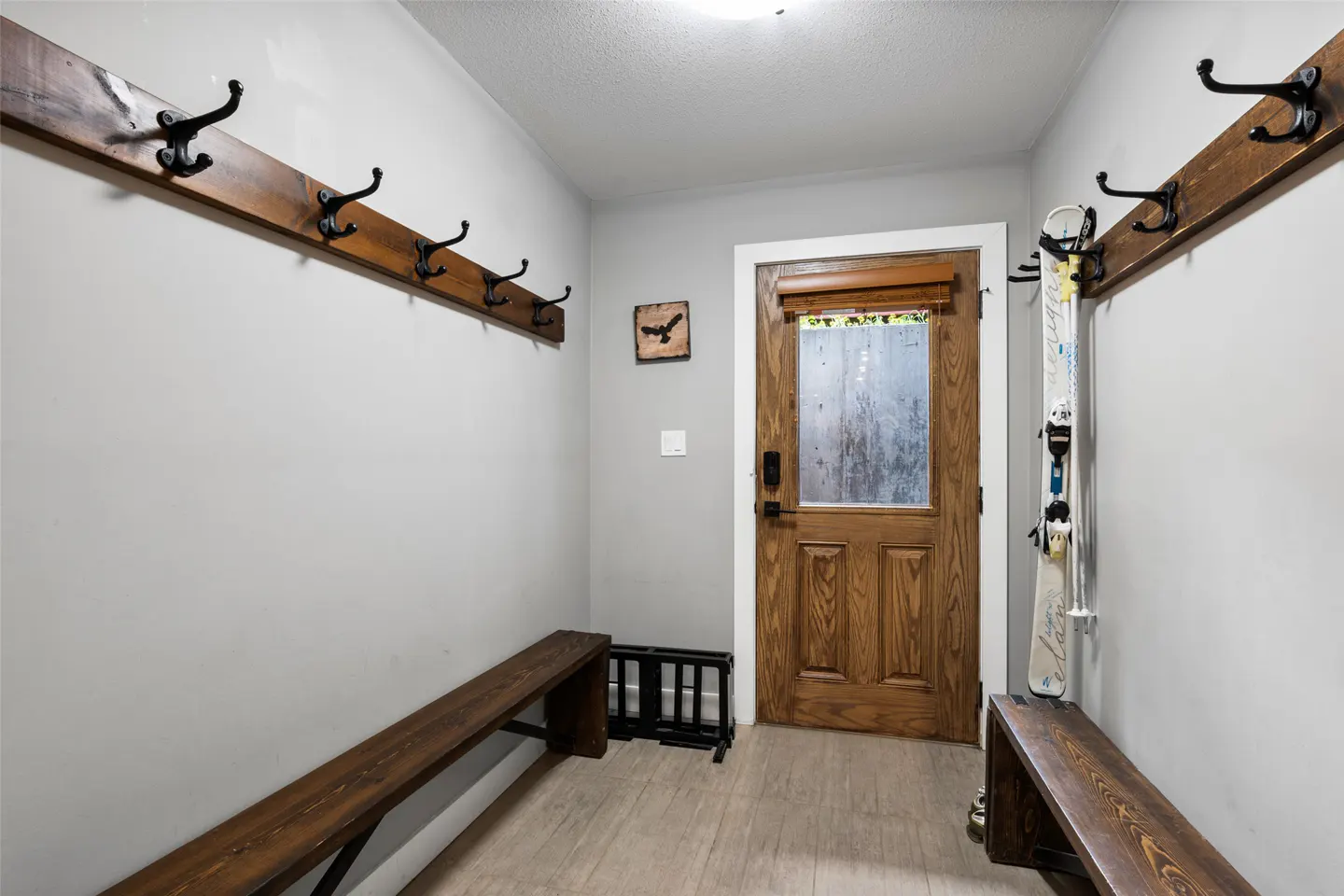 Mudroom with grey walls, wooden benches, wall-mounted coat hooks, and a wooden door with a glass pane.