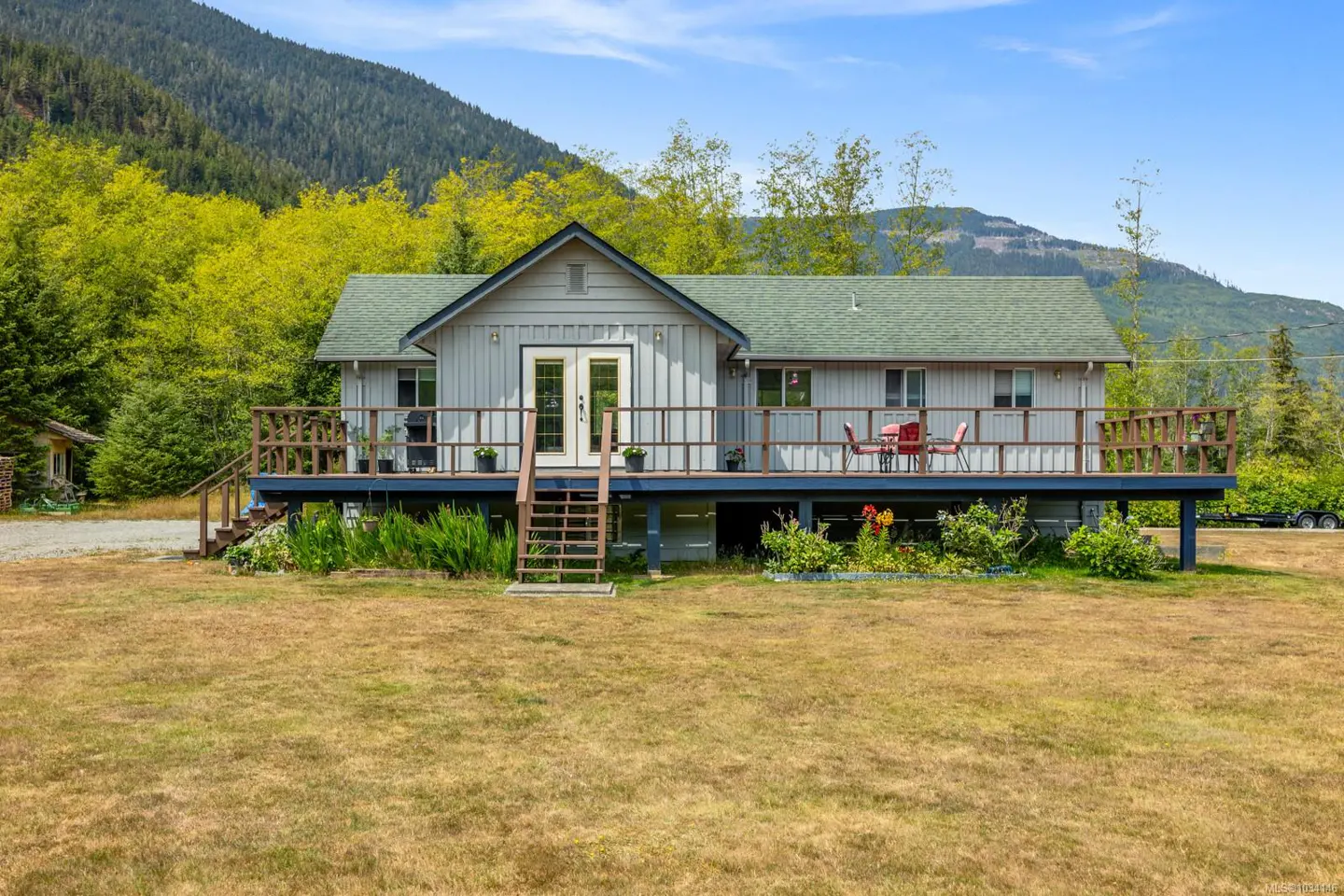 A gray house with a green roof and a large deck, set against a backdrop of mountains and trees.