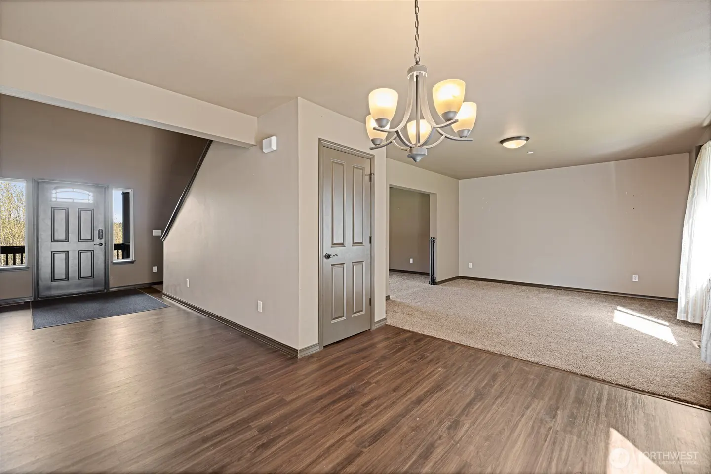 A bright, open-concept home interior with wood floors, beige carpet, and a chandelier. A front door and staircase are visible.