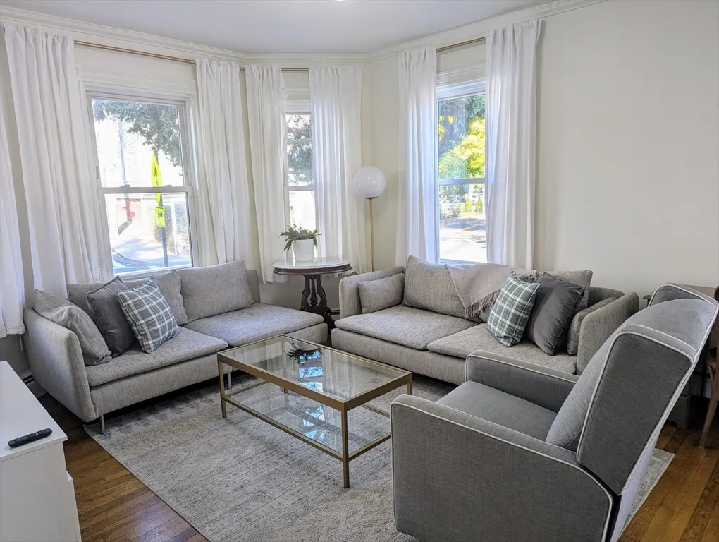 Bright living room with two grey sofas, an armchair, glass coffee table, and white curtains over large windows.