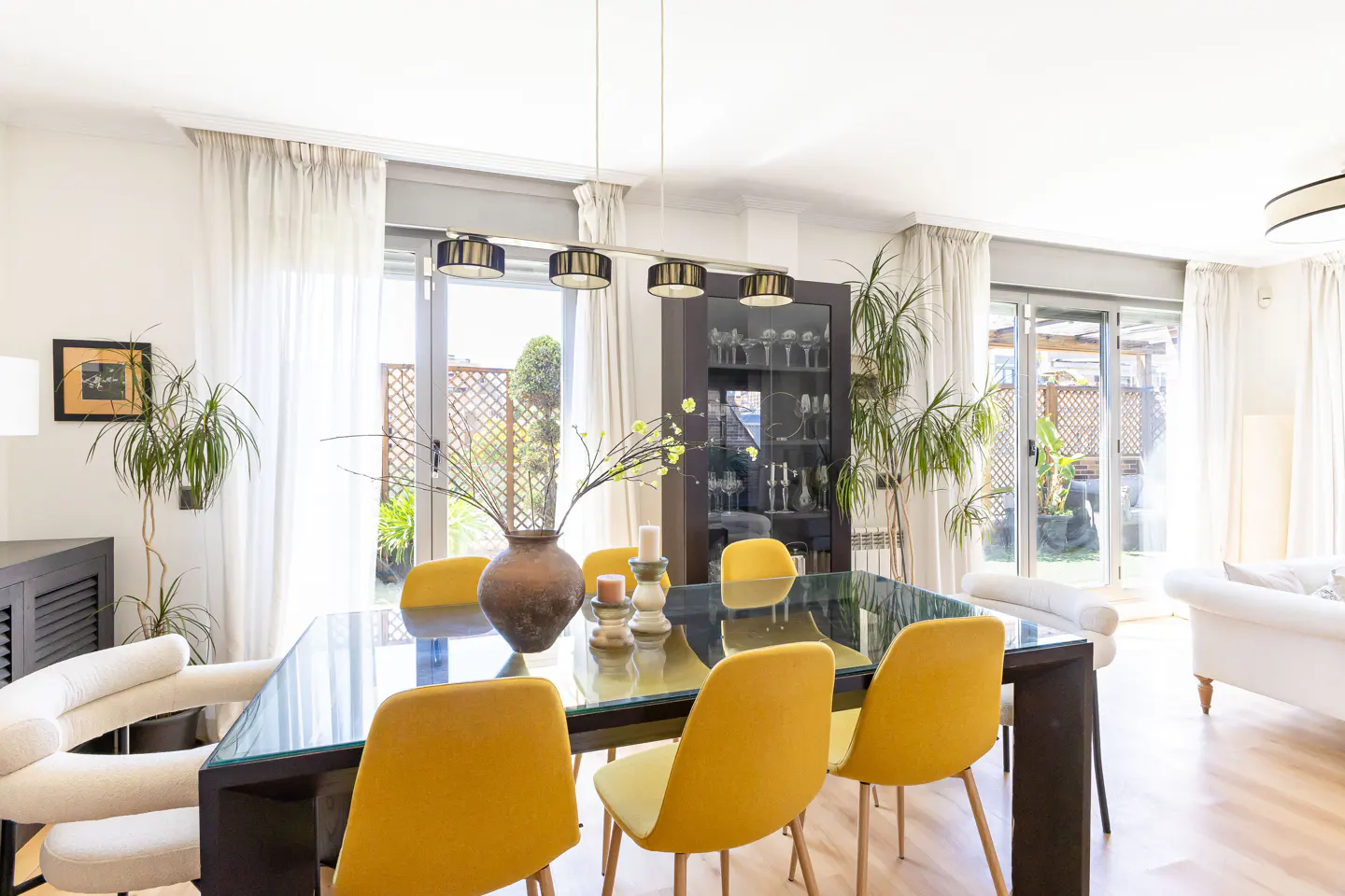 Bright dining room with a glass-topped table, yellow chairs, and a brown vase with flowers. White curtains frame sliding glass doors to a patio.