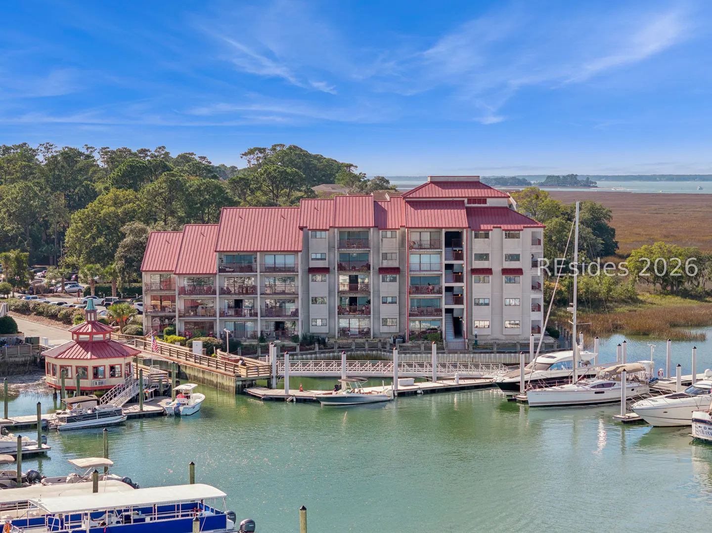 Waterfront condo building with red roof, docks, and boats on a sunny day.