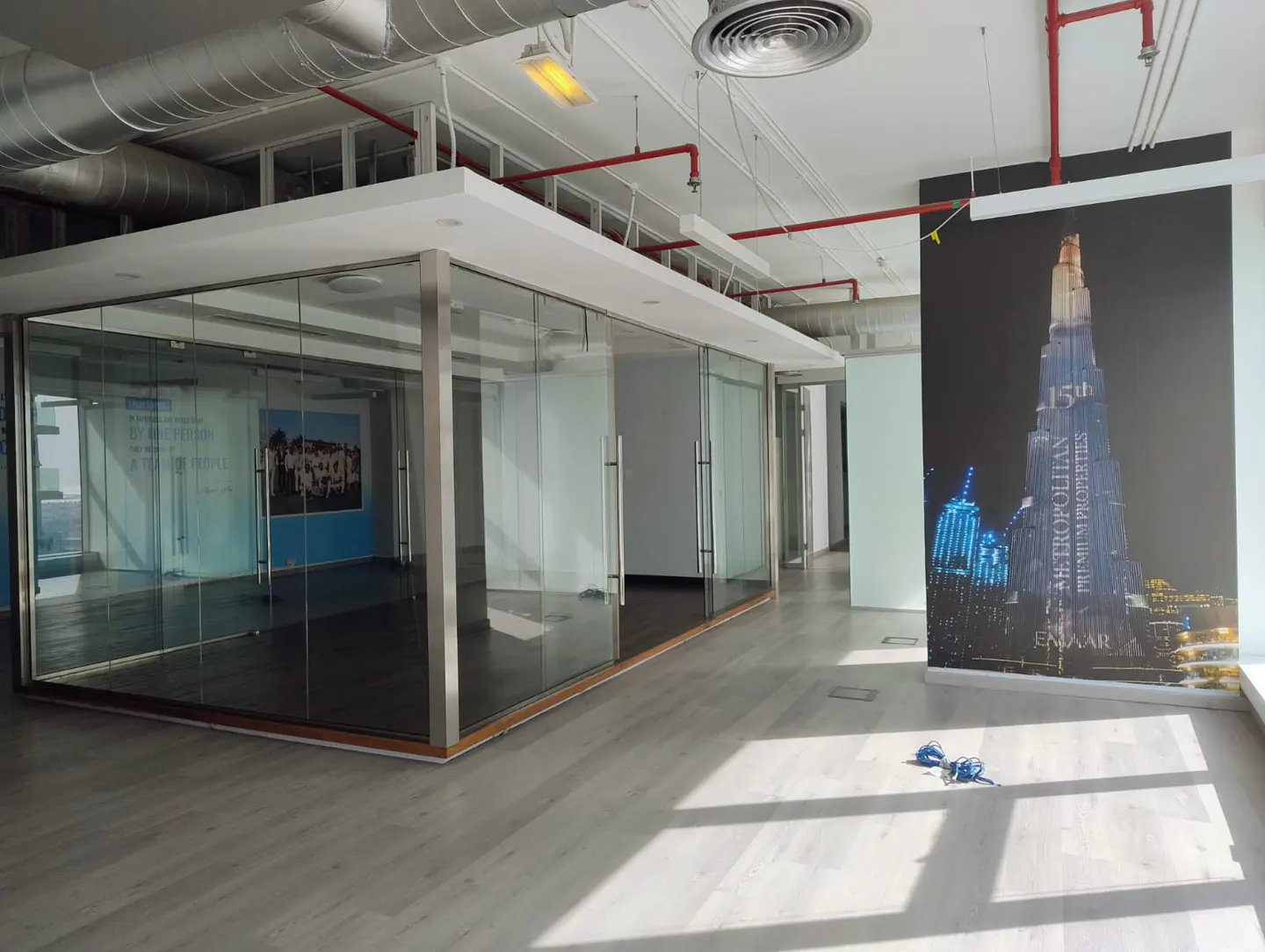 Empty office space with glass-walled rooms, light wood floors, and a large poster of the Burj Khalifa skyscraper.