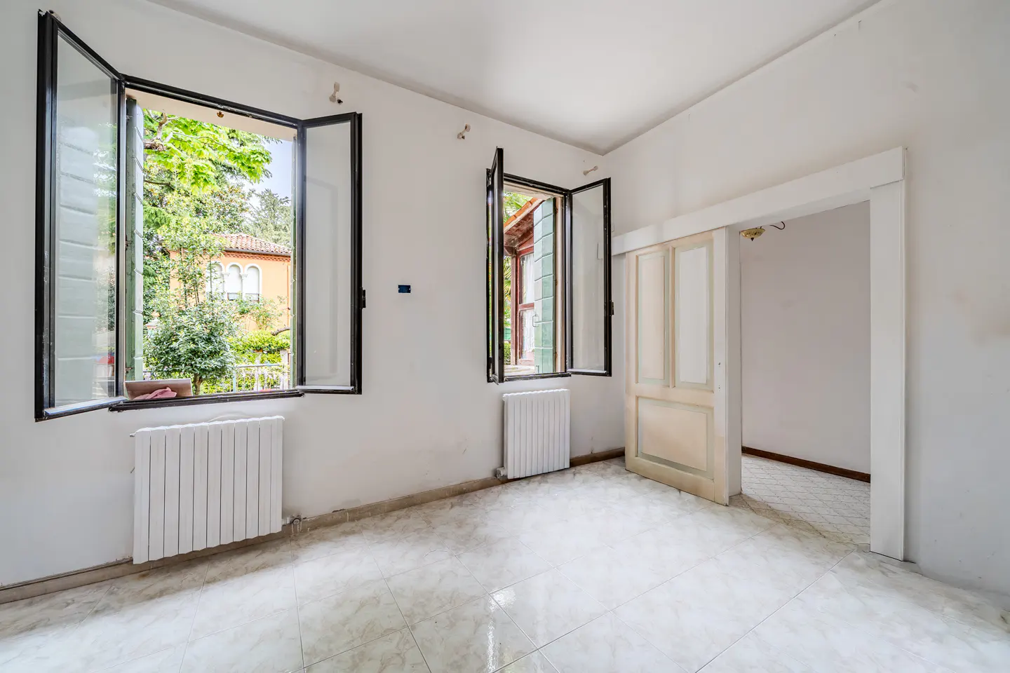 Bright, empty room with white walls, tiled floor, and two open windows showing greenery. A sliding door leads to another room.