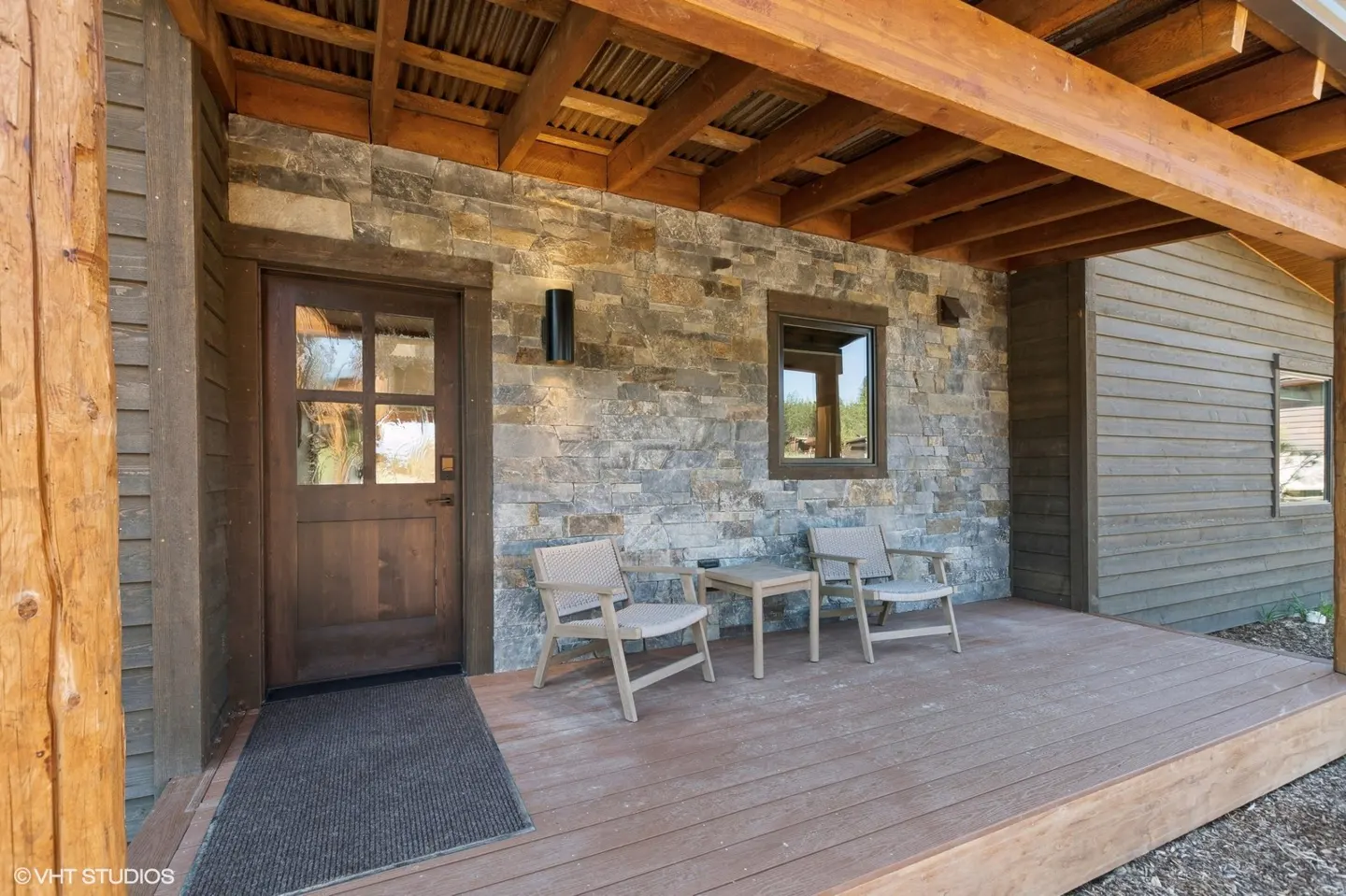 Covered porch with stone wall, wood door, and two chairs with a small table on a wood deck.