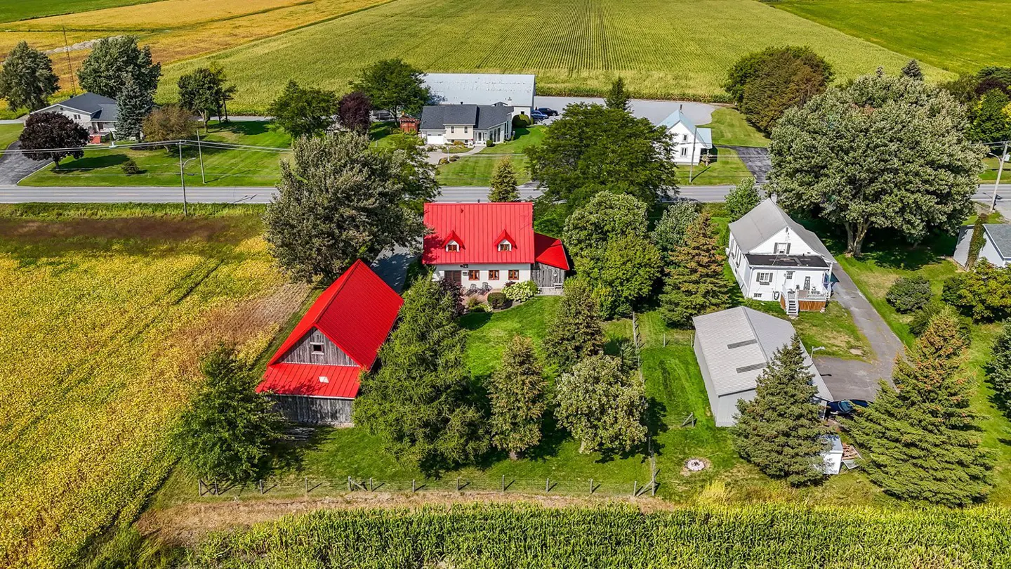 Aerial view of a white farmhouse with a red roof and a red barn, surrounded by green trees and yellow and green fields.