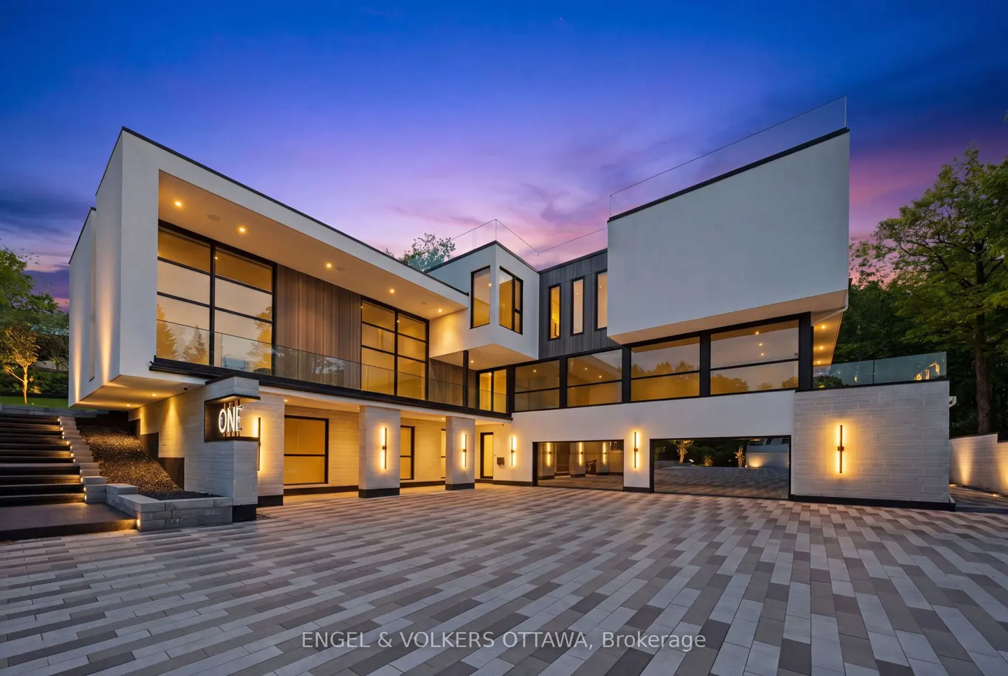 Modern luxury home with white exterior, large windows, and illuminated stone driveway at dusk under a purple sunset sky.