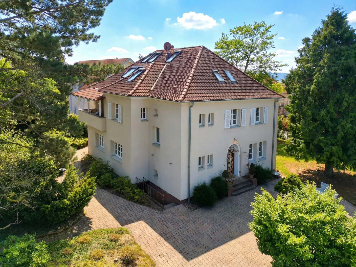 Two-story beige house with a red tile roof and white shutters, surrounded by green trees and a brick driveway.