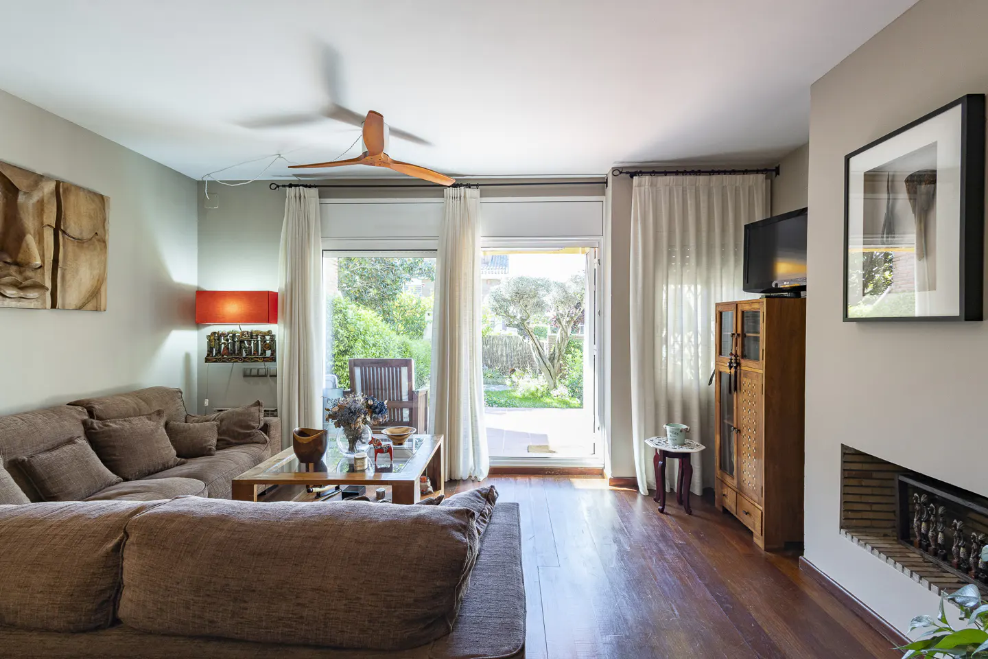 Living room with brown sofa, wood floors, and garden view through glass doors. A ceiling fan and artwork add character.