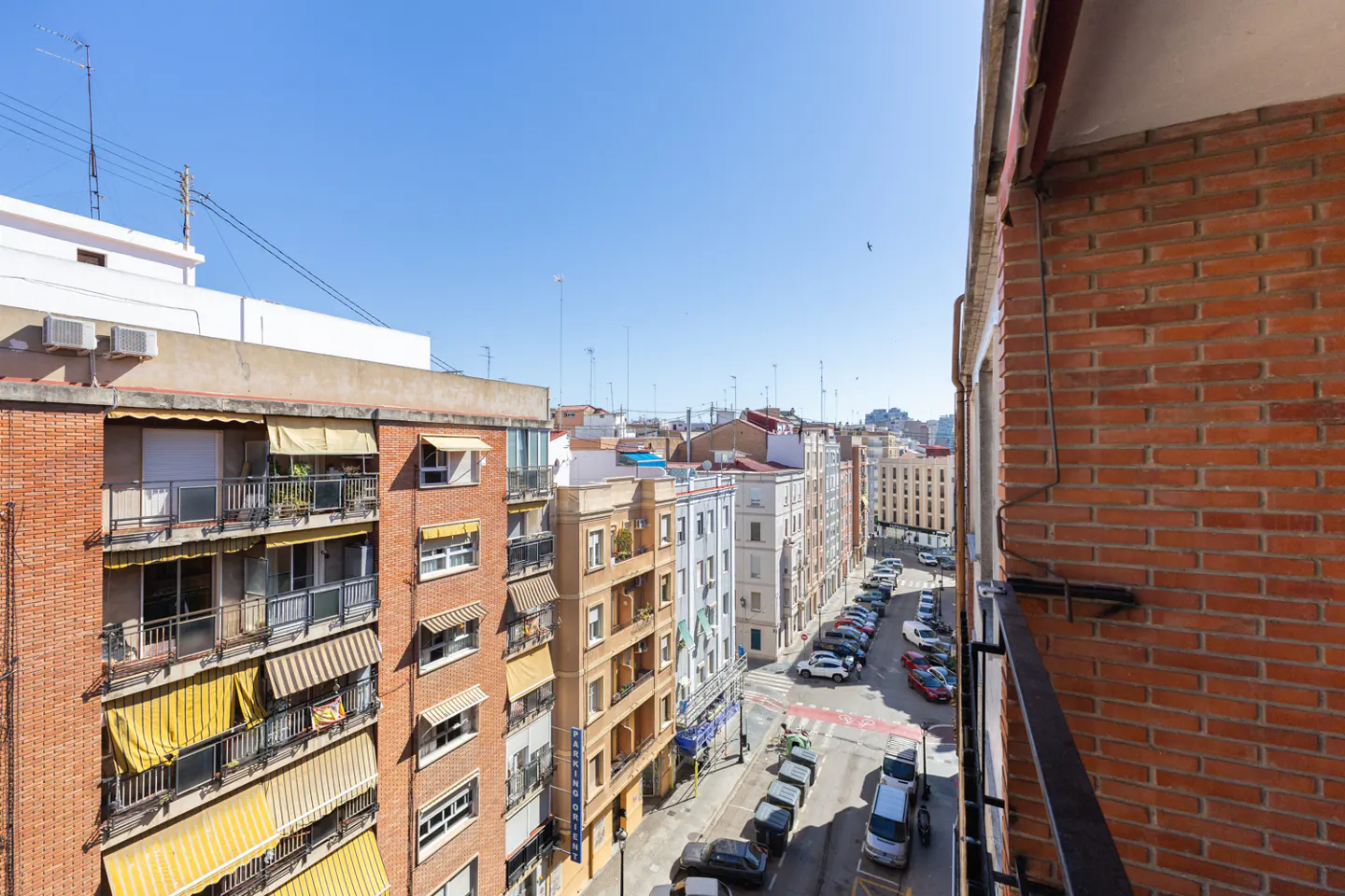 View from a balcony overlooking a city street with parked cars and buildings under a clear blue sky.