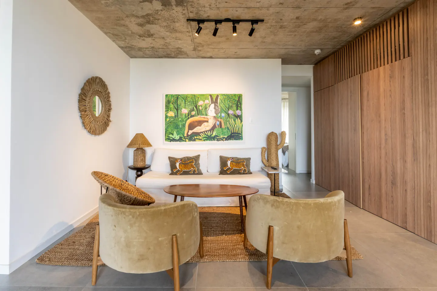 Modern living room with two beige armchairs, a white sofa, a wooden coffee table, and a concrete ceiling with track lights.