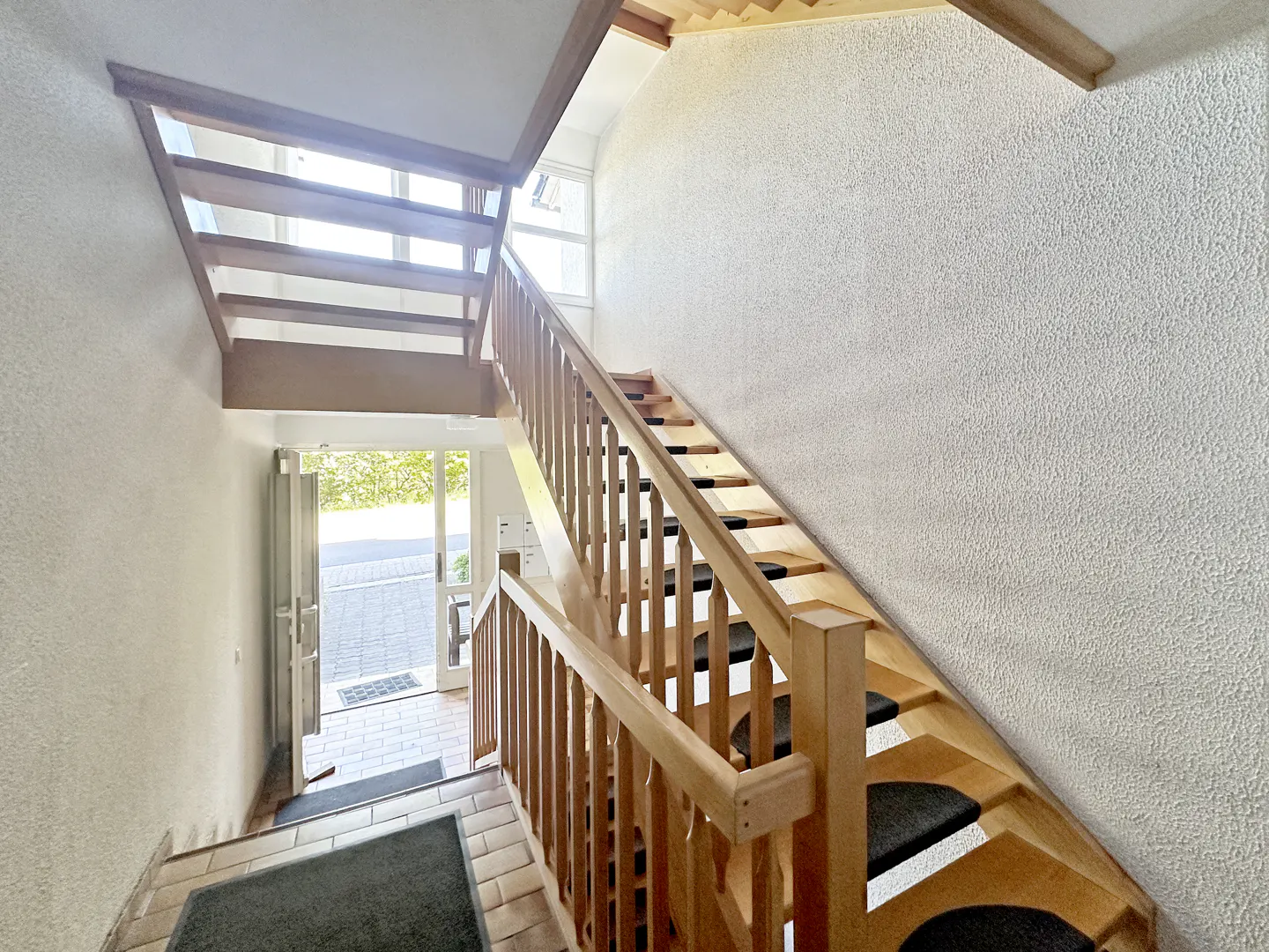 Interior view of a staircase with wooden banister and black treads, leading to an open door with natural light.
