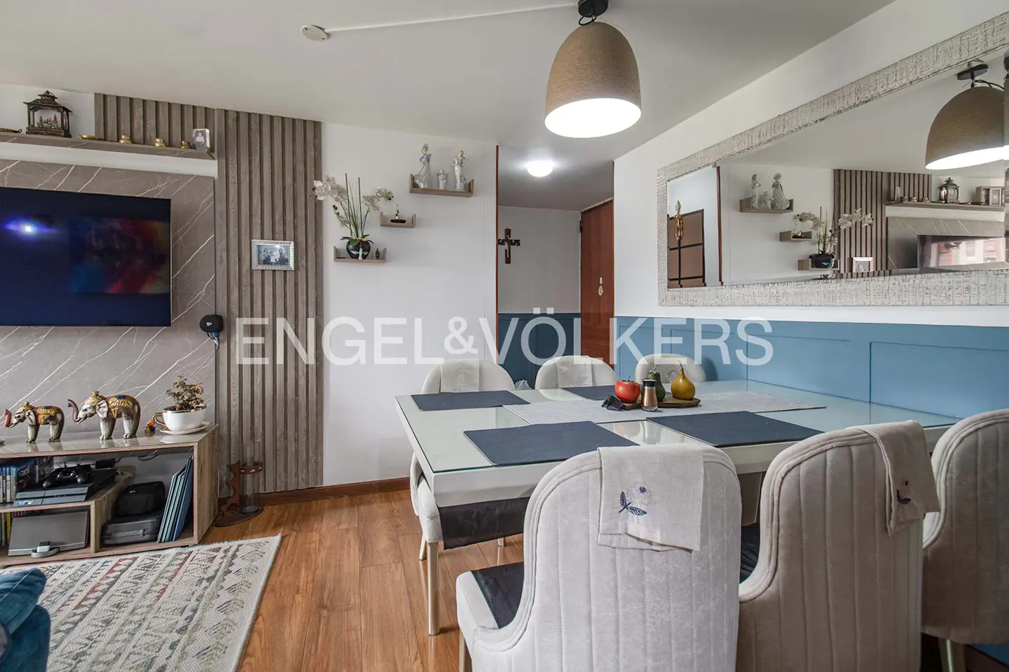 Living room with a glass dining table, upholstered chairs, TV, and decorative shelves. The walls are white and wood paneled.