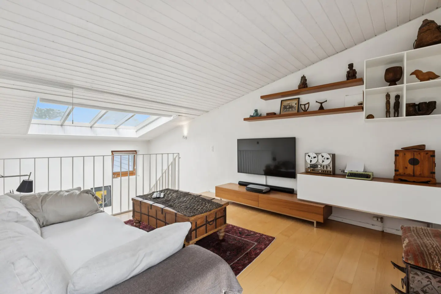 Bright living room with white walls, wood floors, and a white ceiling. A large TV is mounted on the wall above a wooden console. A skylight is visible.