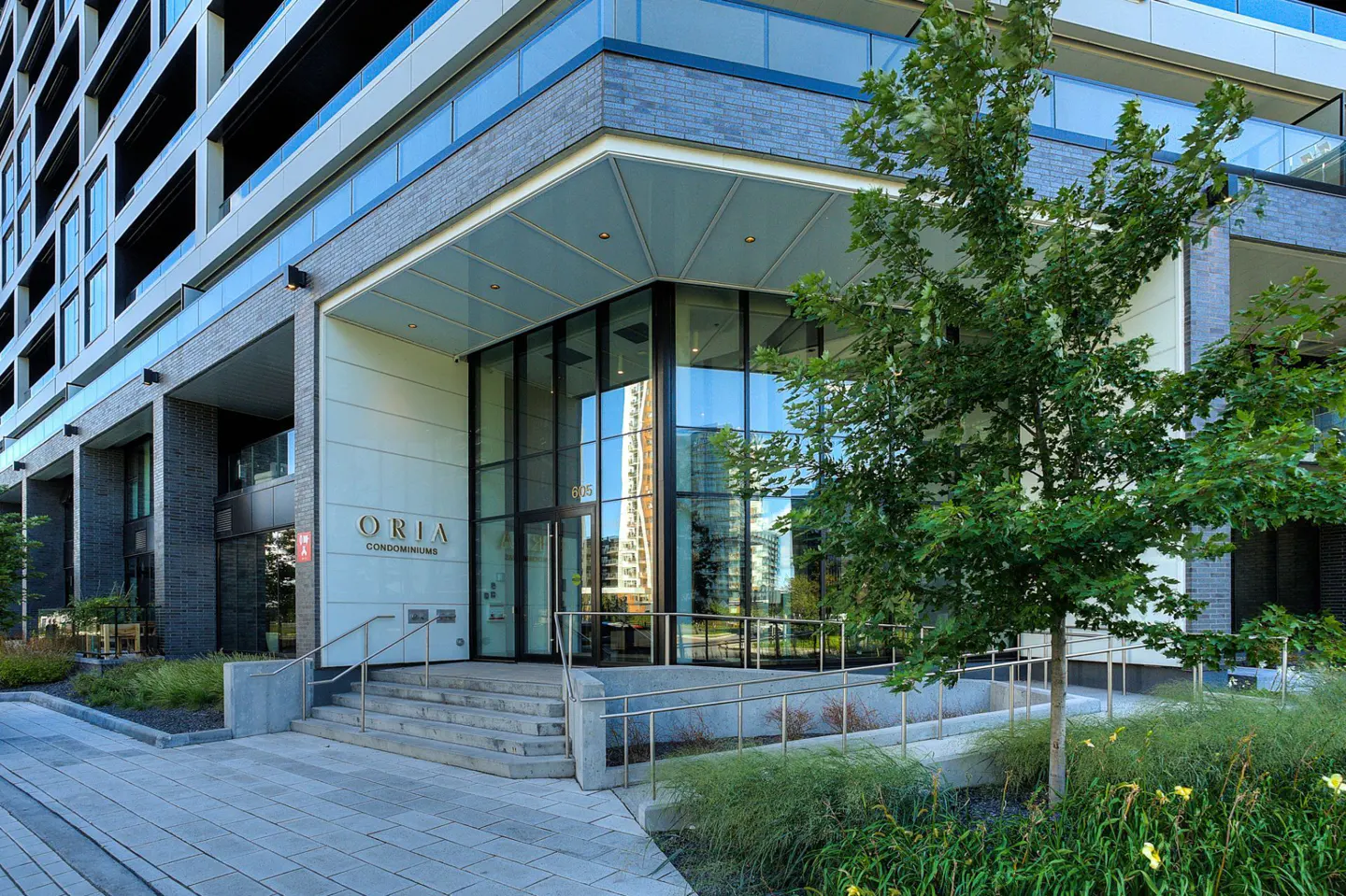 Exterior of Oria Condominiums with glass entrance, steps, and landscaping. The building is modern with gray brick and glass balconies.