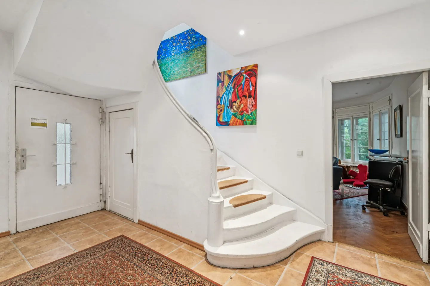 Bright foyer with white walls, tile floor, and a curved staircase. Colorful artwork adorns the walls. A glimpse into a room with a red chair is visible.