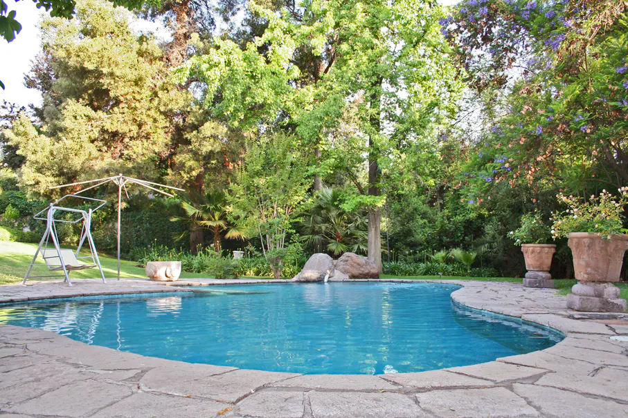 A kidney-shaped pool with blue water is surrounded by a stone patio and lush green trees. A swing set sits on the grass nearby.