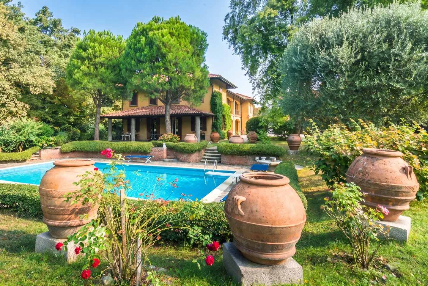 A yellow house with a red tile roof overlooks a blue pool. Large terracotta pots decorate the green lawn.