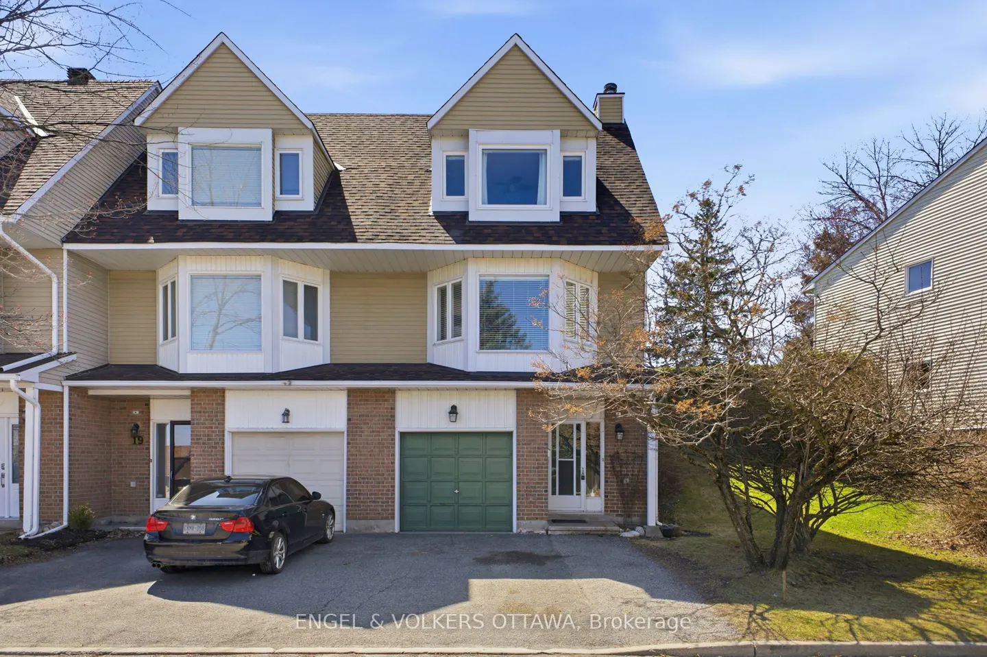 A two-story tan townhouse with brick accents, a green garage door, and a black car parked in the driveway on a sunny day.