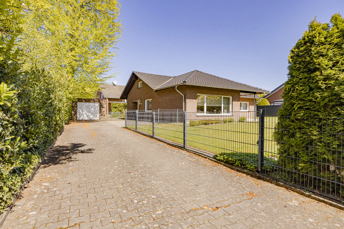 Exterior of a one-story brick house with a dark roof, a metal fence, and a paved driveway on a sunny day.