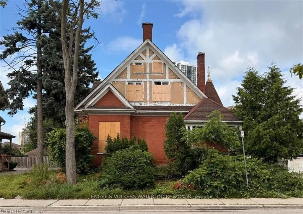 A two-story red brick house with boarded-up windows and overgrown bushes in front. Tall trees frame the house against a cloudy sky.