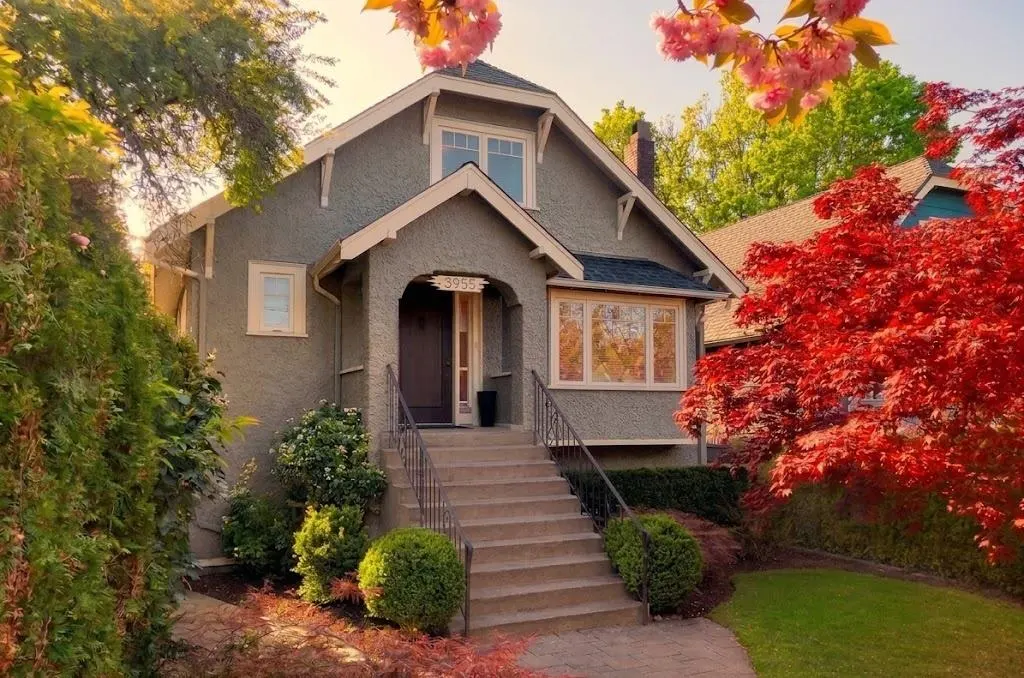 A two-story, gray house with a dark roof, surrounded by colorful trees and bushes. A set of stairs leads to the front door.